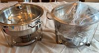 Two stainless steel round chafing dishes on stands with lids and fuel holders visible