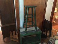 Green painted wooden stool standing on green painted cabinet shelf against a wood door, showing whole pieces including stool legs and cabinet open shelf.