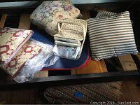 View showing two blue striped pillows, floral pillow, packaged quilted pillow covers, folded linens, woven basket, and braided rugs underneath