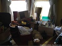 Photo showing multiple boxes on a table containing stained glass pieces, shells, bags of small items, and other miscellaneous craft materials in a room with two curtained windows.