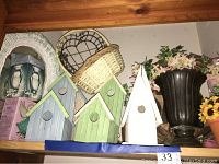 Photo showing four wooden bird houses painted blue and green with circular entrances, a small white wicker framed mirror, wicker basket and artificial flowers nearby.