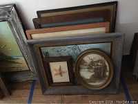 Photo showing all three framed wall art pieces leaned against a wall on the floor. Largest is a beach scene with a gray distressed frame, mid-sized scenic oval painting with gold frame, and small rectangular starfish frame.
