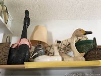 Lamp, black wooden duck sculpture with red scarf, white wooden duck, tan wooden swan or goose, and craft boxes on a shelf.