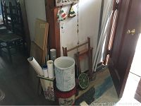 Photo showing vintage wooden stool with canvas seat, metal containers, rolled posters, small chair and other vintage decorative items clustered in a corner.