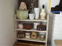 Overall view of painted wooden shelf with assorted pottery items arranged on three shelves.