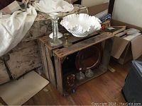 Photo of vintage glassware lot on and inside rustic wooden crate, showing variety of candle holders, white milk glass bowl, carnival glass plate, and blue glass plate.