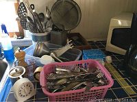 Kitchen counter area with assorted kitchen utensils such as forks, spoons, knives in pink basket, various kitchen tools including strainers, graters, slotted spoons, tongs, timers, mechanical kitchen timer, pepper grinder, and a microwave visible in background.