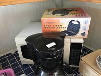 Photo showing white or light-colored Sunbeam microwave oven with keypad and control panel, Toastmaster waffle baker box behind, and Hamilton Beach black coffee maker with visible top lid labeled 'Press to Close'.