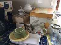Wide view of assorted serving pieces including boxed large gold jar, teapots, stoneware dishes, and fine china cups and saucers.