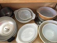 Wood drawer containing stacked white and cream Pyrex bowls and metal bakeware pans with black handles and ventilation holes.