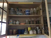 Photo of cabinet shelf with assorted glassware including clear stemmed glasses, blue patterned glasses, and ceramic mugs in various colors.