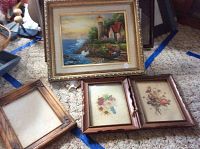 Framed painting of lighthouse with ocean and flowers on a carpeted floor with other items surrounding