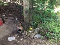 Photo showing assorted metal yard decor including clay chiminea pot, metal basket, and metal stake on soil by wooden post.