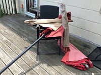 Two outdoor patio umbrellas with beige and red canopies laid on a deck, alongside a folding chair with a dark woven design.