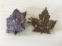 Two WWII Maple Leaf Canadian military cap badges, one dark metal, one lighter metal, placed side by side on a white surface.