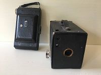 Two vintage folding cameras side by side, one upright and one showing front lens and bellows extended.