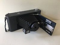 Camera with front door open showing folding bellows, lens, and shutter mechanism.