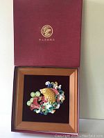 Full view of the tri-color fish art plaque in wooden frame displayed inside the open red box with gold emblem and Chinese characters on lid.
