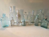 All twelve vintage collectible bottles in various sizes and colors, including three aqua green bottles, arranged in a single line on a white surface against a gray background.