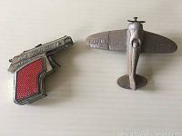 Two vintage toys displayed side by side on white surface: metal airplane marked Pursuit Ship and small metal toy pistol with red textured grip.