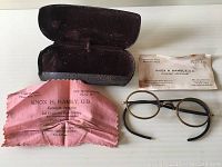Photo of the antique reading glasses, the pink cleaning cloth with Knox H. Hambly's details and the optometrist card, alongside the open eyeglass case.