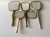 Front view of five vintage hand held vanity mirrors with varying shapes and metal finishes, including one missing its mirror glass.