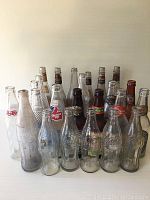 Photo showing 24 beverage bottles of various brands and types arranged in rows featuring clear and brown glass bottles