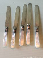 Five silver plate knives with iridescent mother of pearl handles laid flat on a surface.