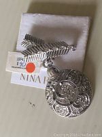 Photo shows new Nina Ricci brooch with a detailed silver-tone round pendant decorated with swirls and clear rhinestones. Attached feather-shaped element also has rhinestone accents. Original tag visible.