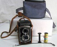 Group shot of camera in leather case, film roll, empty spool, cable release, fabric camera bag