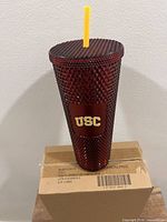 Front view of one red studded Starbucks USC tumbler with yellow straw on cardboard box