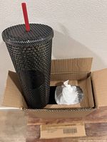 Tumbler inside shipping box showing studded design, red straw, logo medallion