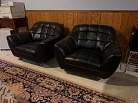 Front view of both black faux leather lounge chairs showing tufted backs, seats, arms, and casters