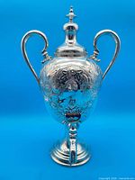 Front view showing bright-cut engravings, double handles, finial lid, pedestal base