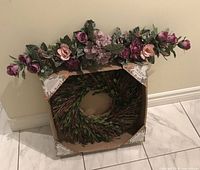 Wreath inside original cardboard packaging below faux floral swag leaning against wall