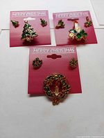 Front view of three carded Christmas jewellery sets showing tree, candle, and wreath designs