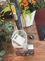 Front view of red and gray cordless upright vacuum with dust bin and charger on floor