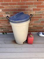 Beige plastic trash can with blue lid, handles and wheels shown against brick wall