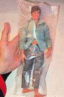 Figure in clear plastic bag held by hand, showing full view, clothing, and damaged leg