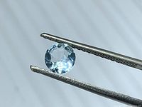 Round faceted aquamarine gemstone held by tweezers, light blue color