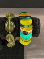 Both bracelets displayed on bracelet stand—chunky multicolor disc bracelet and slim light-green oval bead bracelet