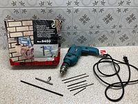 All items laid out: drill, cord, bits, chuck key, depth rod and box