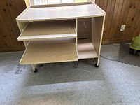 Front view of light oak laminate computer desk showing three pull-out trays, right-side open compartment, and casters