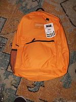 Front view of orange EastWest backpack with tag, showing front zip pocket and mesh side pocket