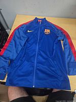 Front view of blue and red Nike FC Barcelona track jacket laid flat