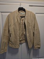 Front view of beige faux leather bomber jacket on hanger showing entire garment, zipper, pockets, sleeves
