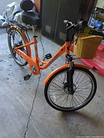 Front three-quarter view showing orange step-through frame, basket, disc brake, fender, kickstand