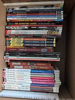 Stack of graphic novels in box showing Kill Shakespeare, DMZ, The Adventure Zone, Angel Catbird, Stitch, Champions, Saga, Skyward, Test and more