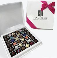 Closed white Anna Stubbe gift box with magenta ribbon alongside open box showing 36 assorted chocolates