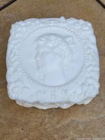 Top view of square white milk glass trinket box lid with raised female cameo and floral relief